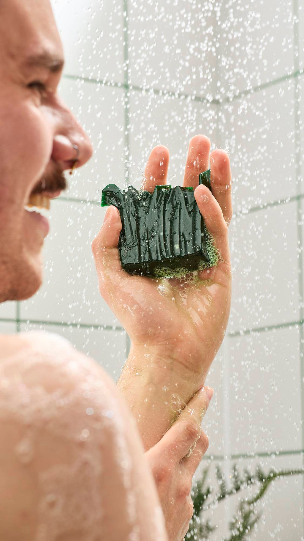A smiling person in the shower holds a textured, dark green shower gummy soap in their hand. Water sprays around them against a backdrop of white tiles with green grout.