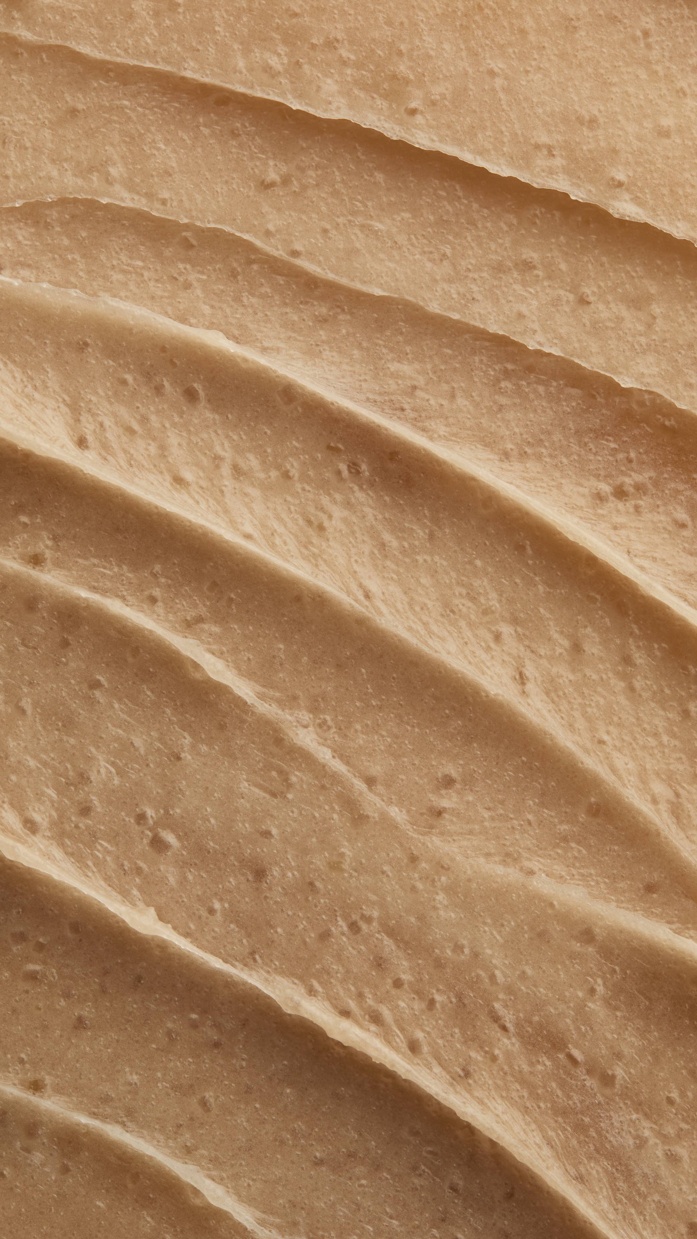 A close-up, full-frame shot displays the texture of the "Crème Brûlée" Body Scrub. The creamy, light brown mixture is spread into thick, wavy ridges, with small exfoliating granules visible throughout the paste.