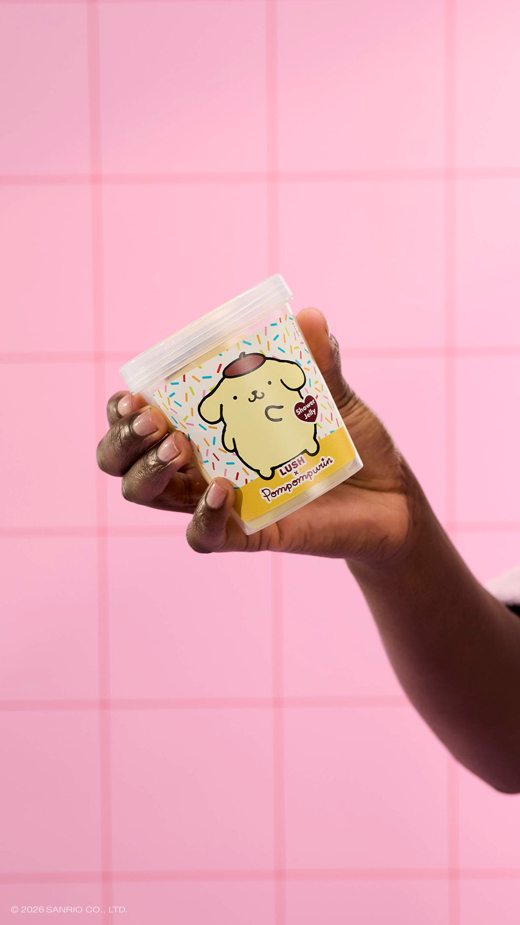 A hand holds a clear plastic tub of Lush x Pompompurin shower jelly, featuring a yellow dog character on the label. The background consists of pink square tiles.