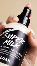 A hand with iridescent manicured nails grips a bottle of "Super Milk" conditioning hair primer. The clear bottle reveals a creamy white liquid inside, while the black label describes the product as an "almond, coconut and oat milk hydrator".