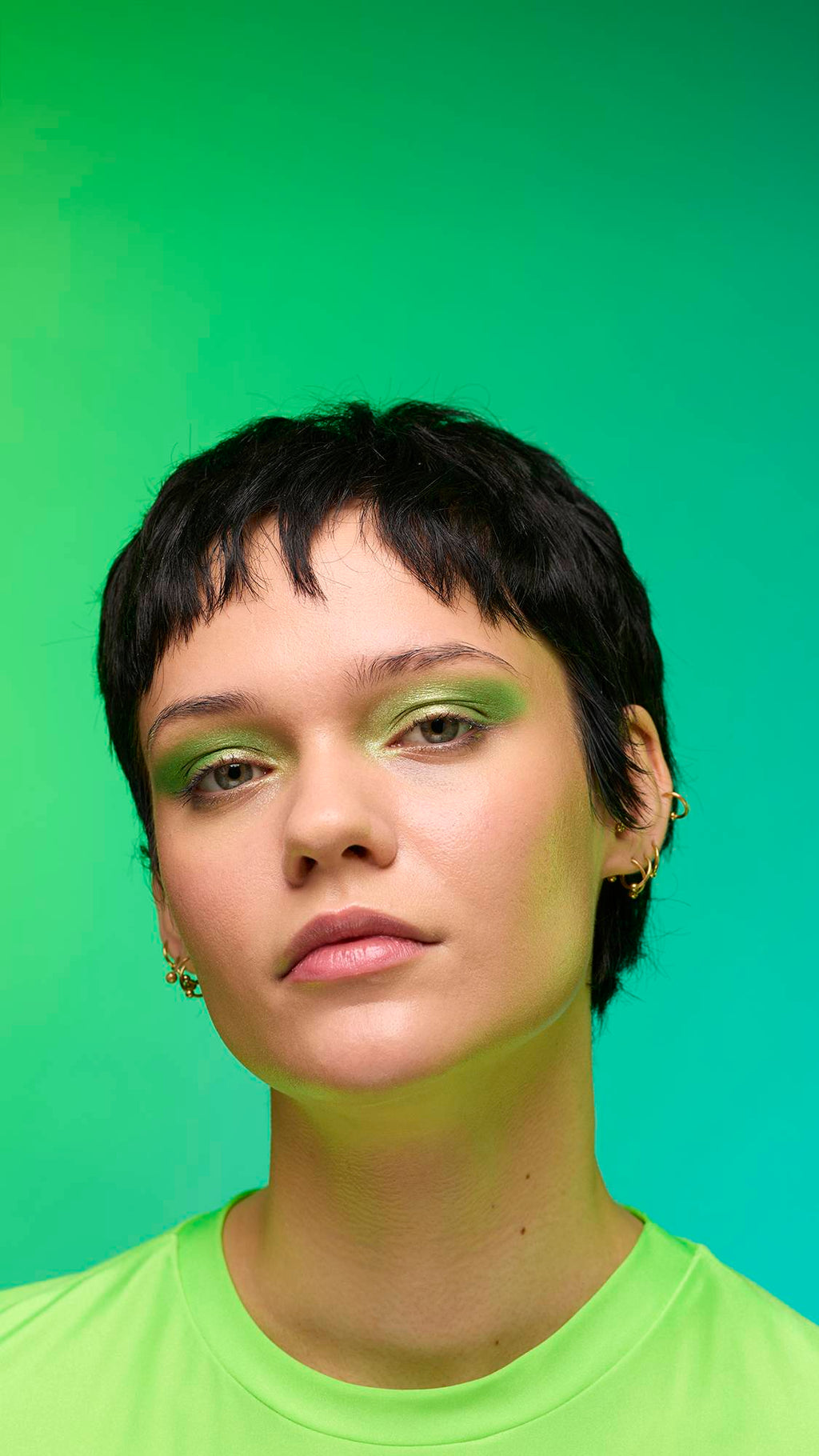 The image shows a close-up of the model's face on a pale blue-to-green gradient background. The model's make-up is done with the focal point being a subtle, jade-green, shimmering eyeshadow made by the Aurora Glitter Crumble.