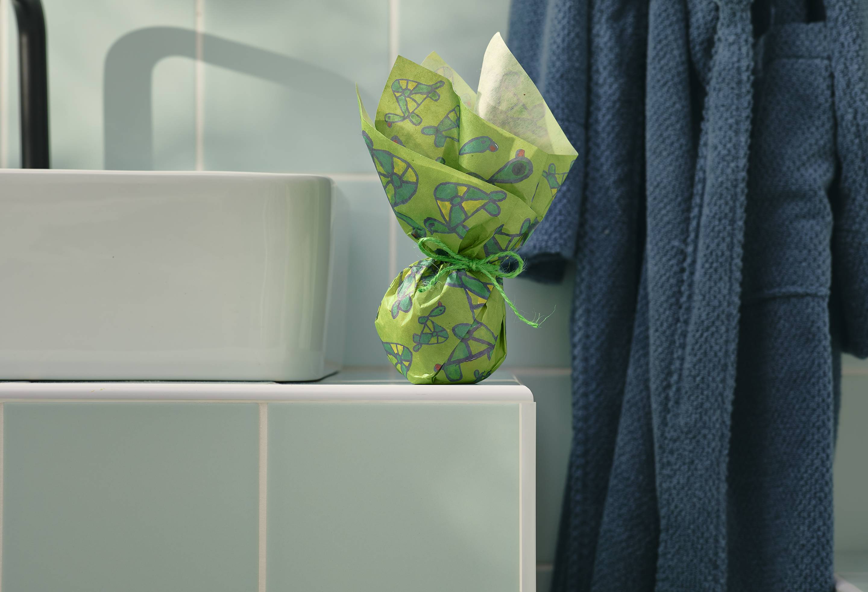 A small, spherical gift, wrapped in green "Turtley Awesome" Lokta paper with a hand-drawn turtle pattern, sits on a tiled bathroom ledge. The paper is gathered at the top and tied with twine. A dressing gown hangs in the background.