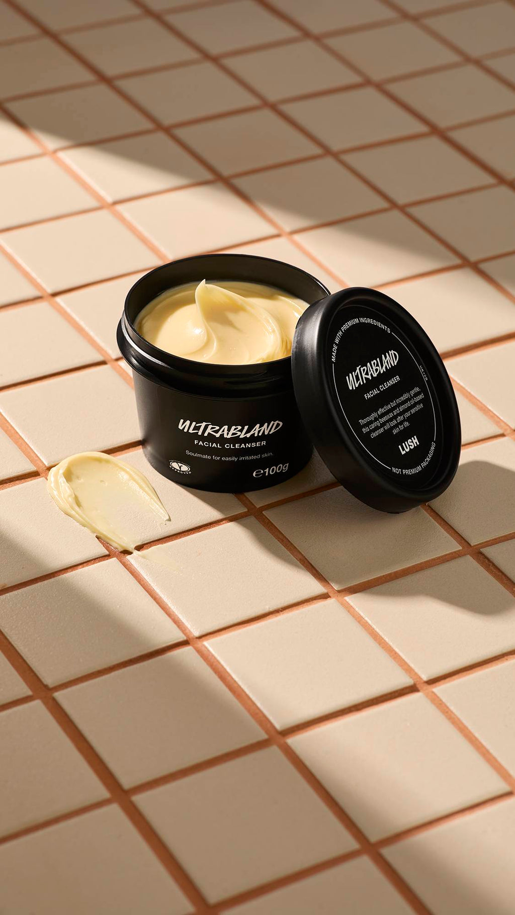 A black pot of Lush Ultrabland Facial Cleanser sits on a beige tiled surface with the lid leaning against it and a smear of the thick yellow cream nearby.